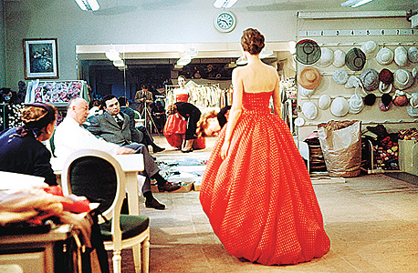 כריסטיאן דיור (משמאל, בחלוק לבן) בוחן שמלת ערב בעיצובו לפני תצוגה (ינואר, 1957)