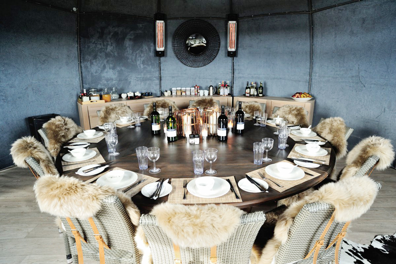  בחדר האוכל מוצב שולחן מעץ אלון שמוקף בכיסאות עטופים בפרווה, שיוצרים מראה כפרי.
