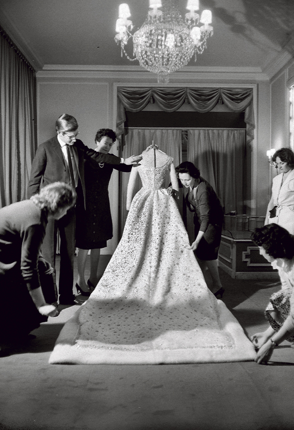 תיקונים אחרונים לשמלת הכלולות של פארה דיבה, שנישאה לשאה הפרסי, בעיצוב איב סאן־לורן (משמאל) לדיור. השמלה משובצת פנינים מלאכותיות, פאייטים וחוטי כסף ומעוטרת בשולי פרווה, הכל בעבודת יד. 1959