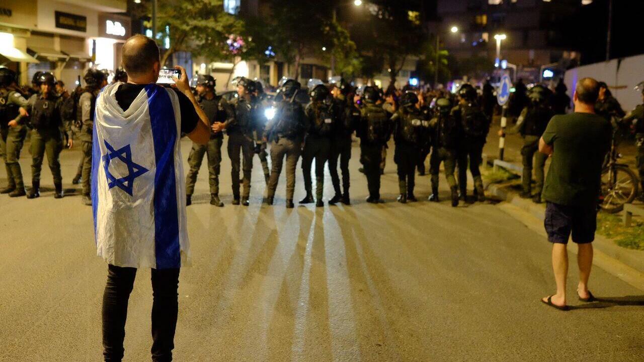 מפגין עם דגל ישראל מול שוטרים שמקיפים את המספרה שבה נמצאת שרה נתניהו בכיכר המדינה בת"א