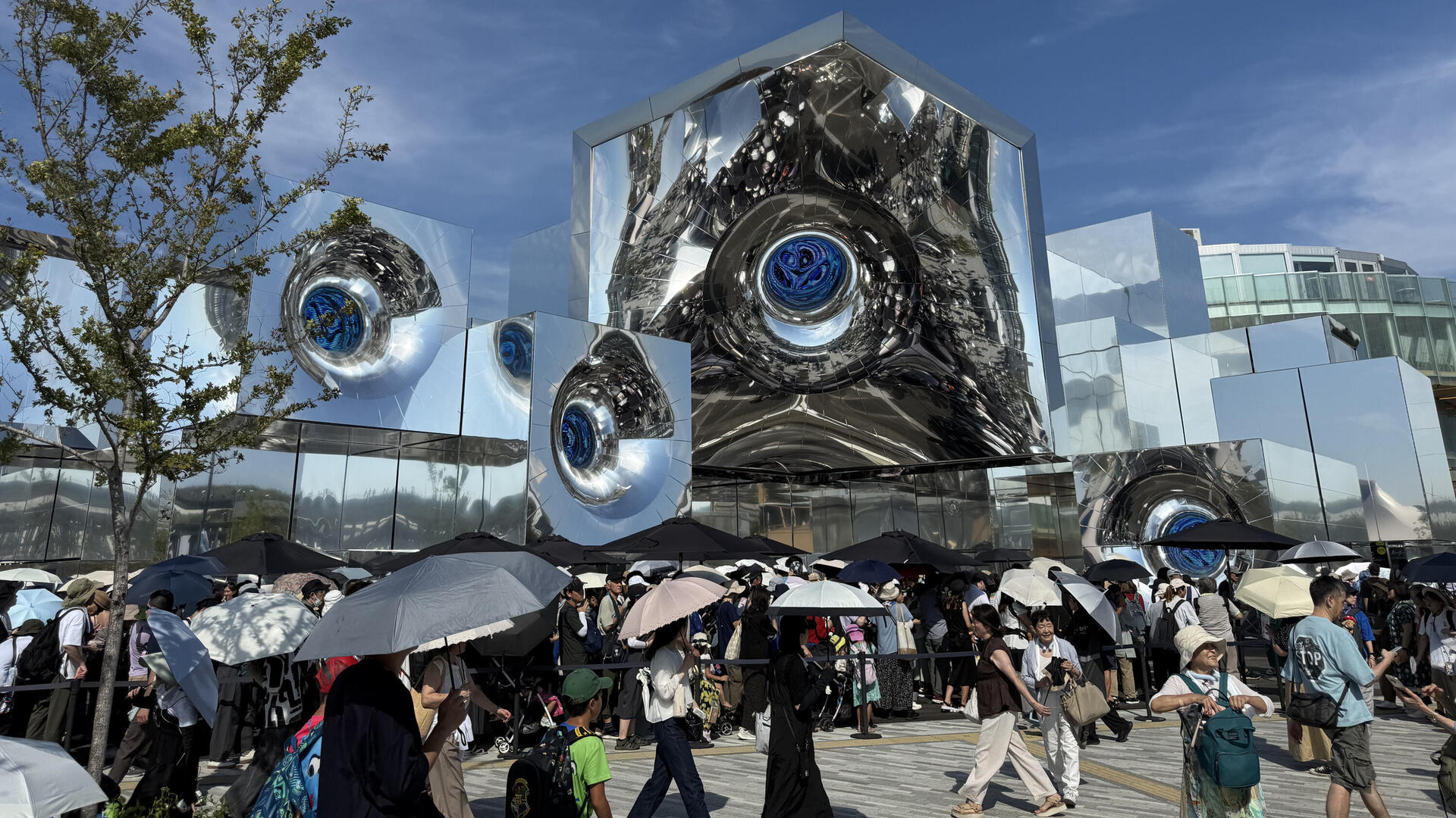 מבקרים באקספו על רקע הביתן של האמן היפני ,Yoichi Ochiai בספטמבר. המארגנים הודיעו שהם שמחים שגם גברים התחילו להסתובב במתחם עם שמשיות, וגם הפעילו ממטרות לצינון. זה לא עזר