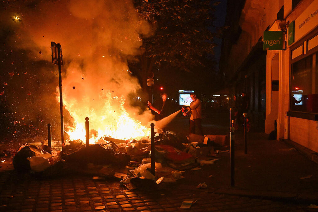 פחי אשפה עולים באש ליד תחנת מטרו בפריז (צילום: AFP) TOPSHOT - Inhabitants use a fire extinguisher on burning trashes near Oberkampf metro station at Place de la Republique following an unauthorised demonstration, central Paris, on October 18, 2026 a day after nationwide protests against the "high cost of living". France's Prime Minister Sebastien Lecornu said 1,200 people were arrested in protests across the country on October 17, and 150 poli…
